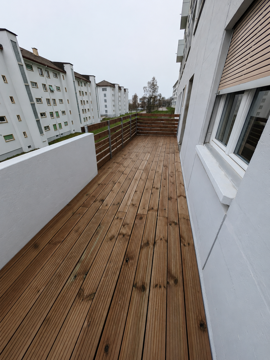 Balcon en bois sur le toit d'un immeuble, avec murs et balustrades blancs sous un ciel couvert.