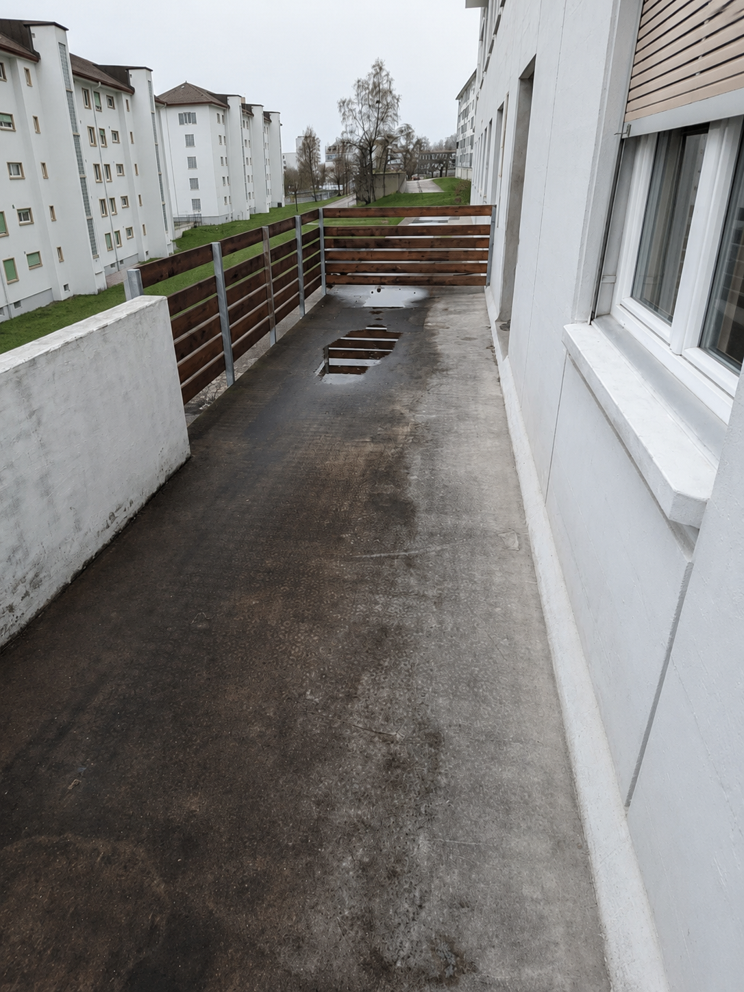 Allée piétonne extérieure étroite en béton longeant des immeubles d'appartements blancs, avec une clôture en bois basse et un revêtement mouillé.