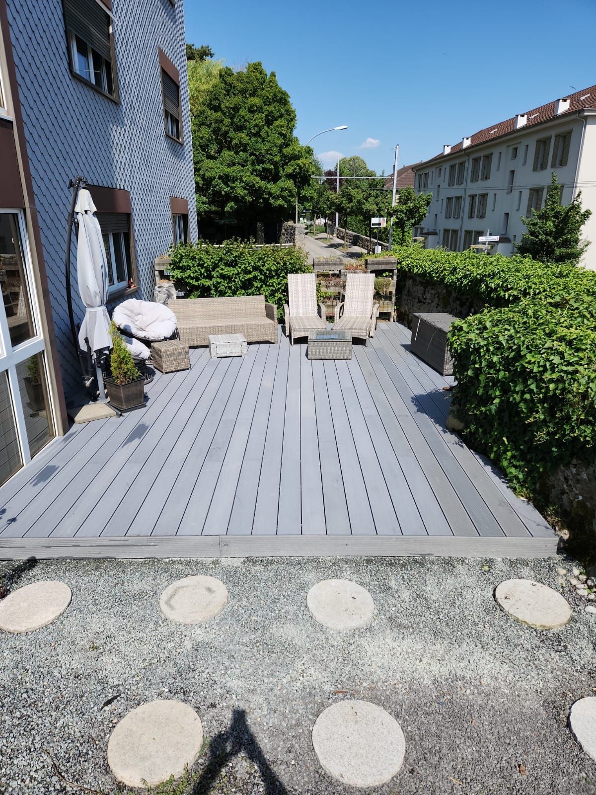 Terrasse grise moderne avec chaises et table de jardin à côté d'une maison bleue, donnant sur un chemin de gravier et un jardin.