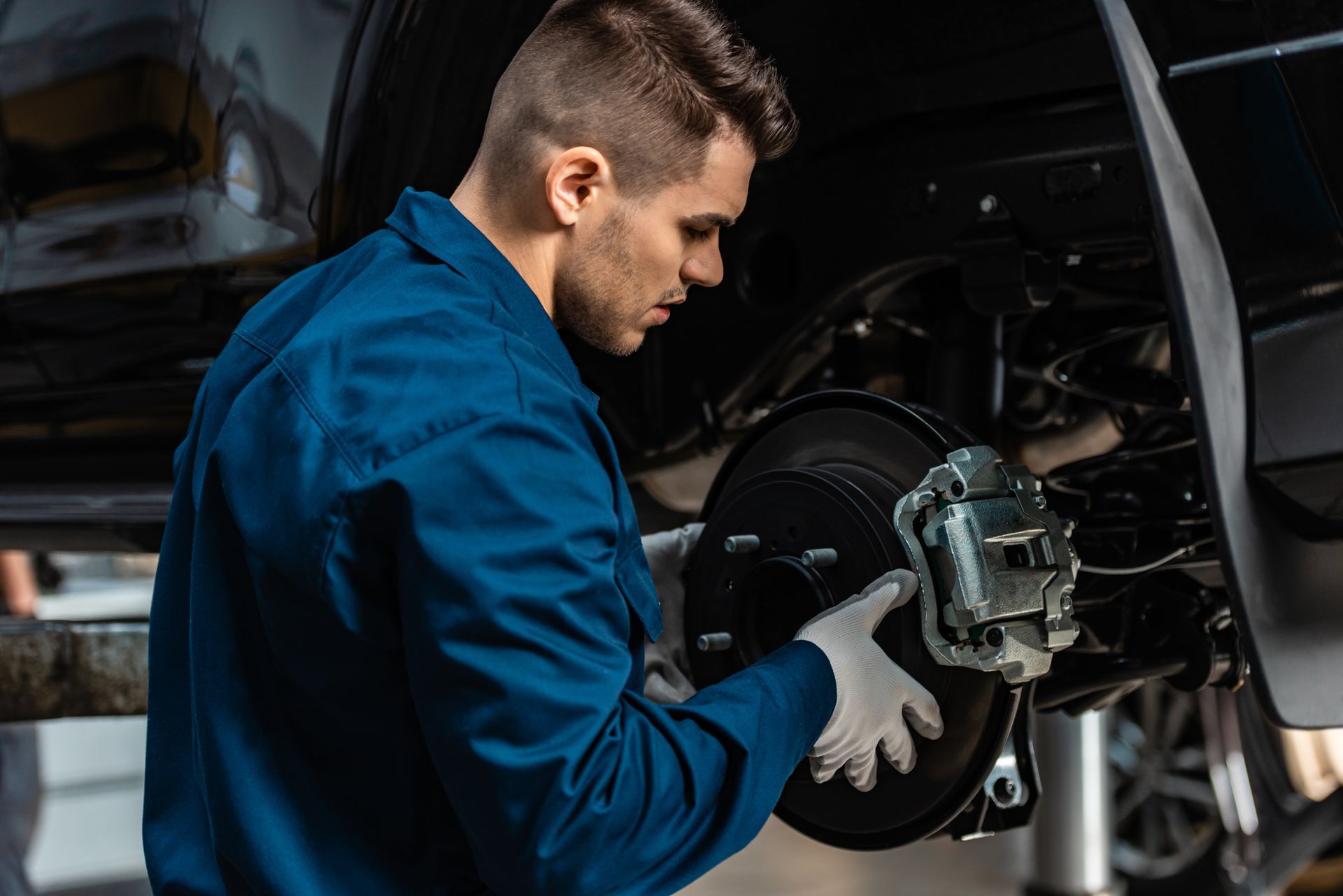 Mechanic in blue uniform, examining car brake system, wearing gloves.