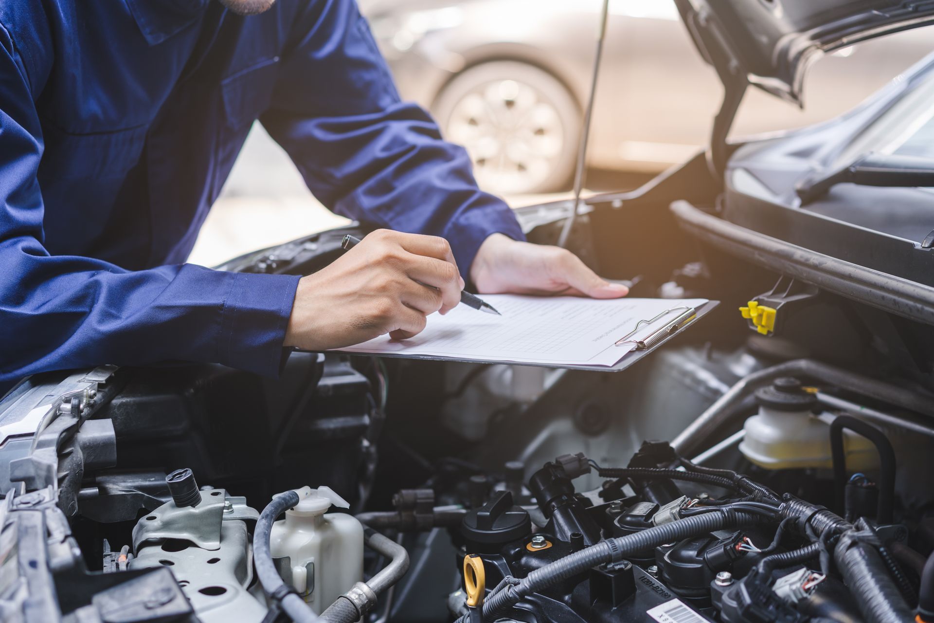 Mechanic in blue overalls inspecting car engine, holding clipboard outdoors.