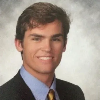 Portrait of a smiling person in a dark suit, blue shirt, and patterned tie against a gray background