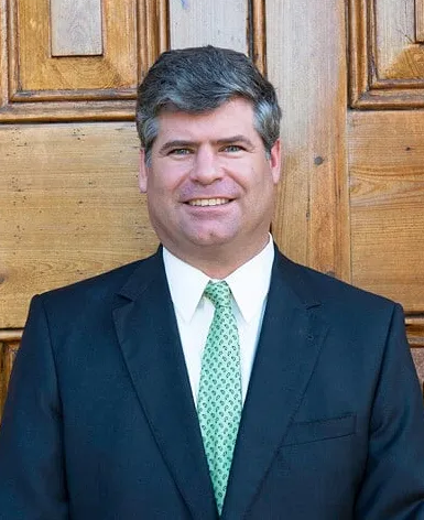 Man in a dark suit and green tie standing in front of wooden double doors, smiling.