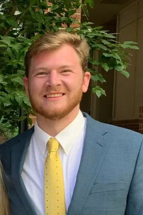 Smiling man in a blue suit with a yellow tie standing outdoors near greenery.