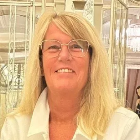 Smiling blonde person wearing glasses and a white collared shirt indoors, with decorative glass background