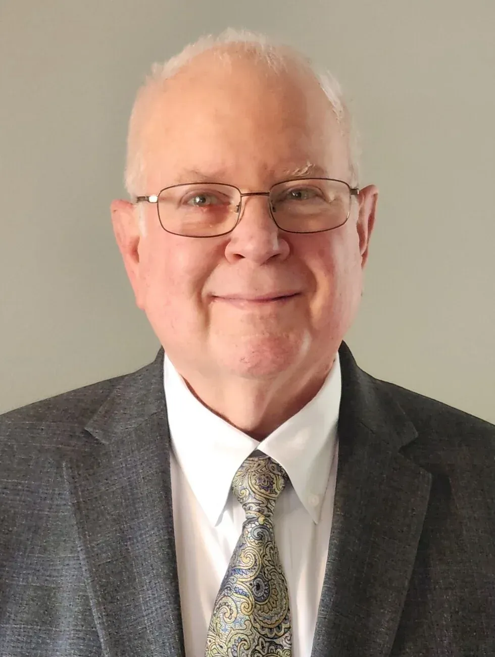 Portrait of a smiling man in a dark suit, white shirt, and patterned tie against a plain gray background