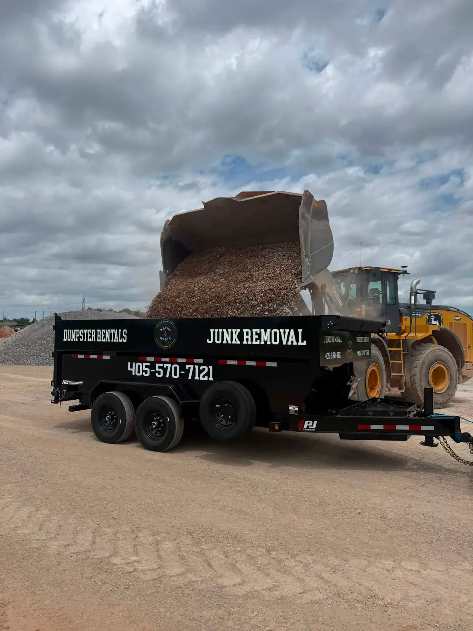 Dump truck unloading dirt into a black junk removal trailer on a gravel lot under cloudy skies
