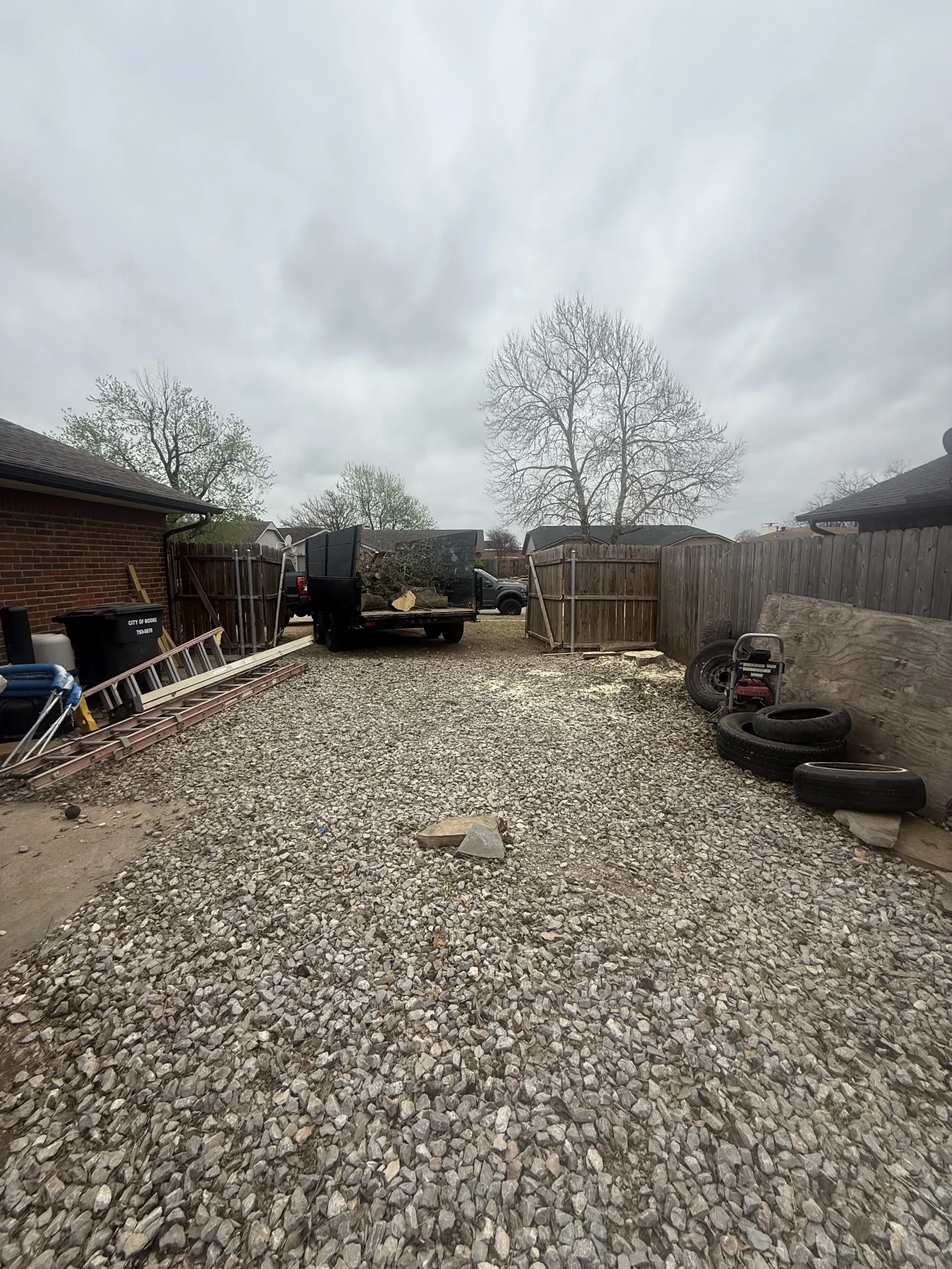 Gravel driveway beside a shed and fence, with tires and a gate under a cloudy sky