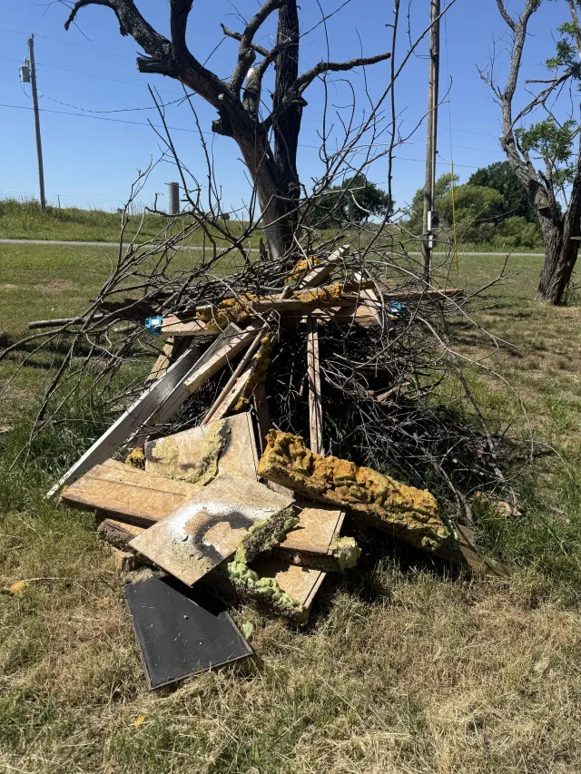 Burned pile of wood and debris in a grassy field under a clear blue sky
