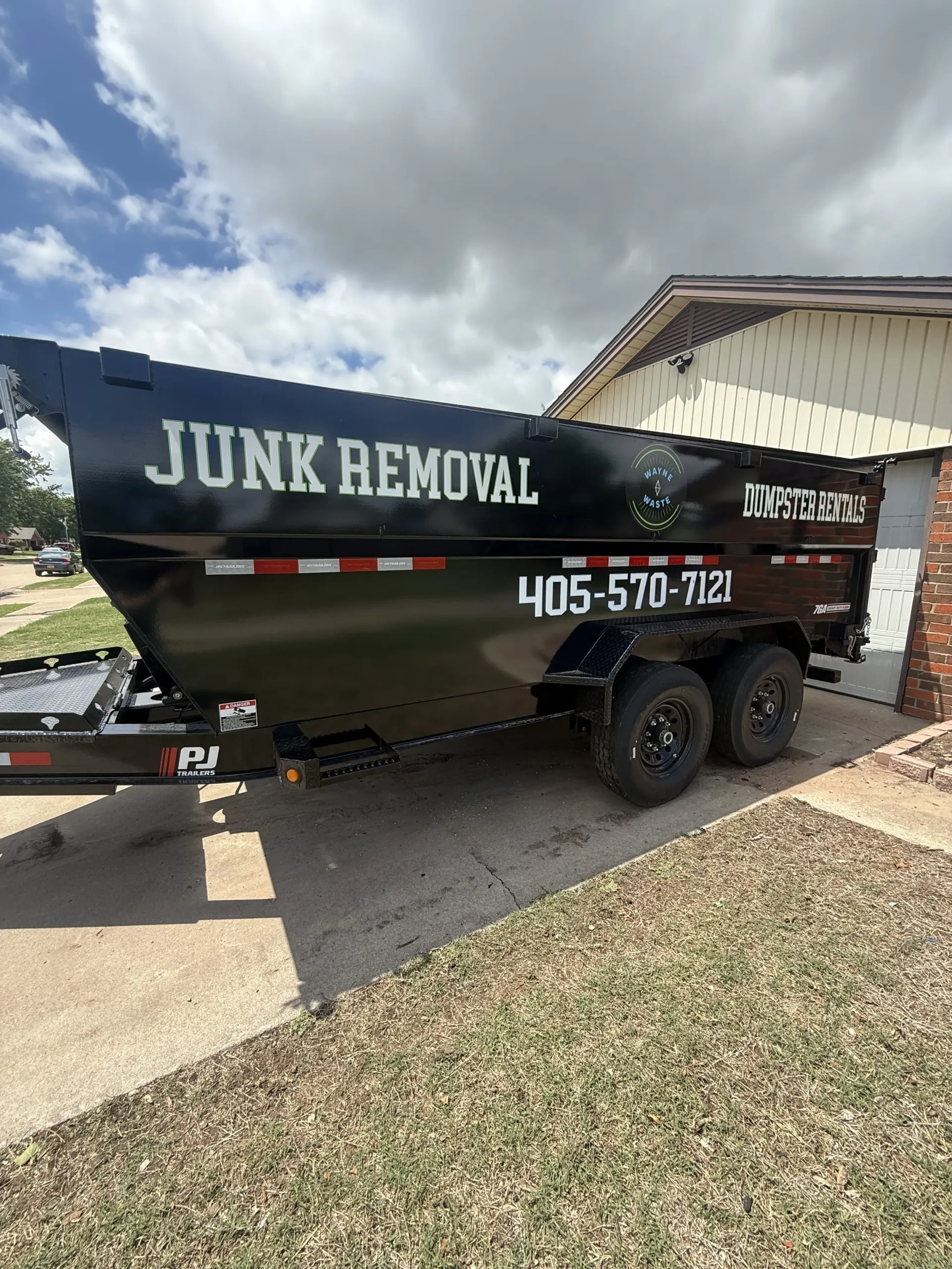 Black junk removal trailer parked in a driveway under a partly cloudy sky.