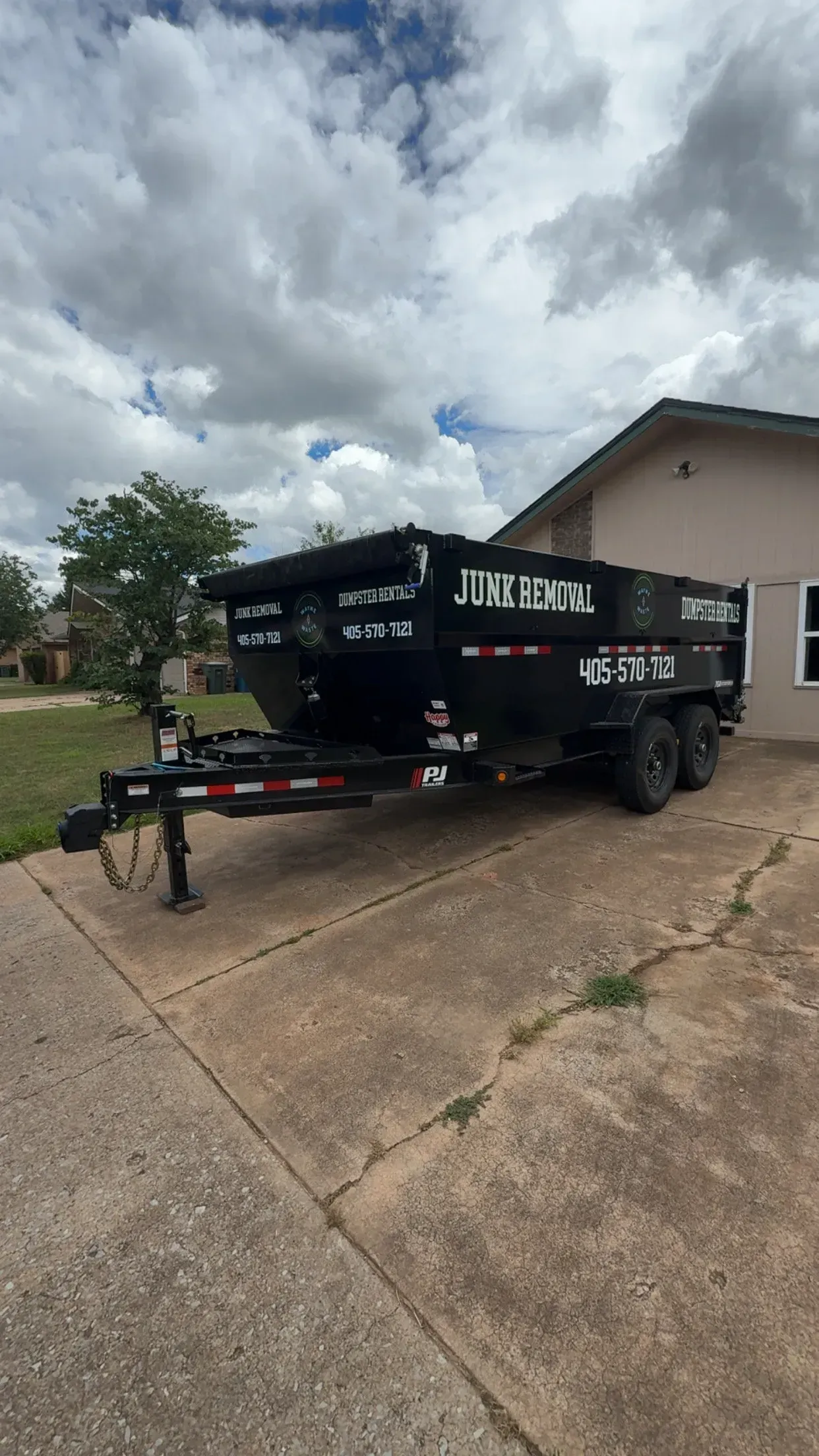 Black dump trailer parked on a driveway beside a house under a cloudy sky.