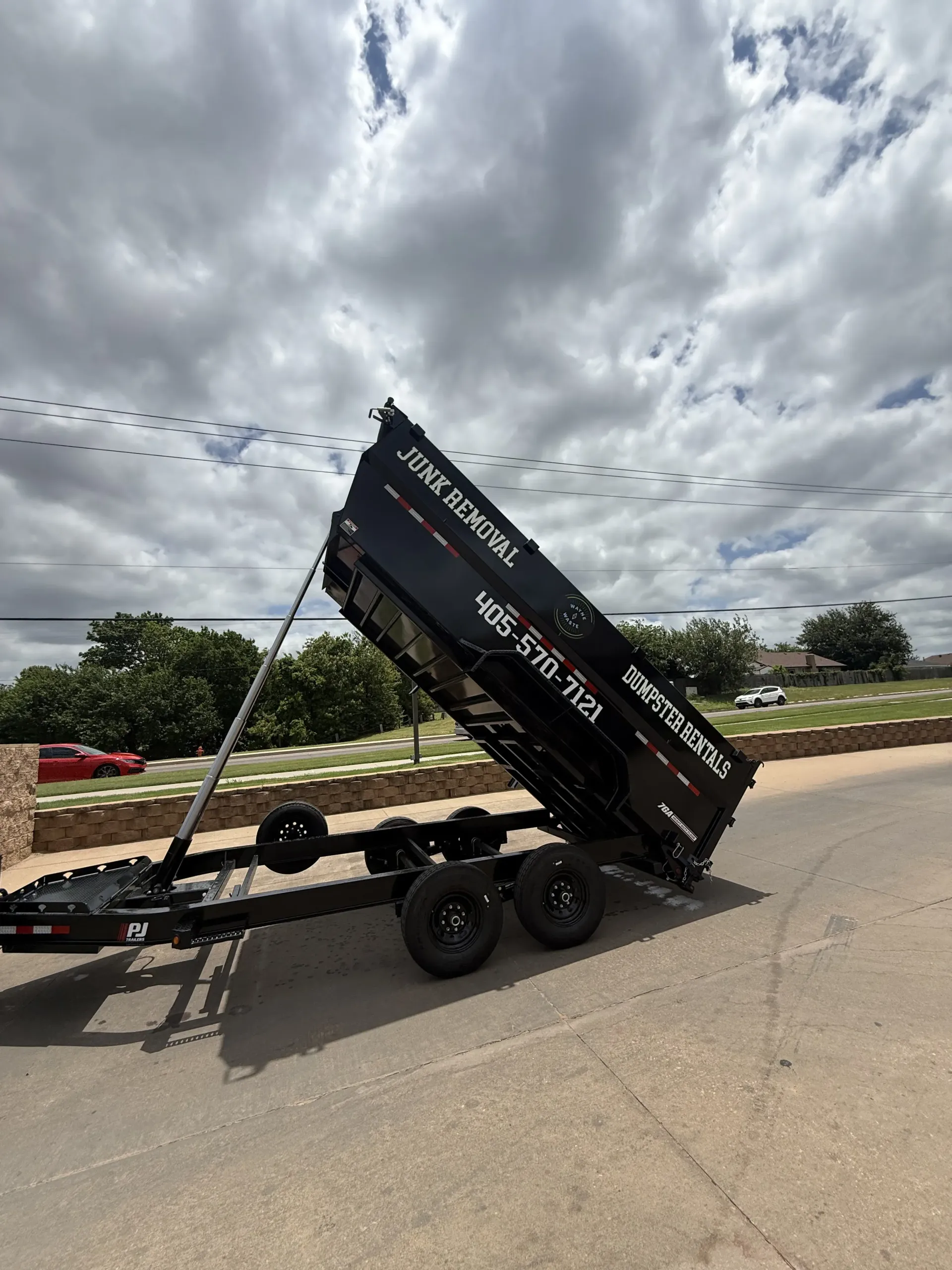 Black dump trailer tipped steeply on a road under a cloudy sky.
