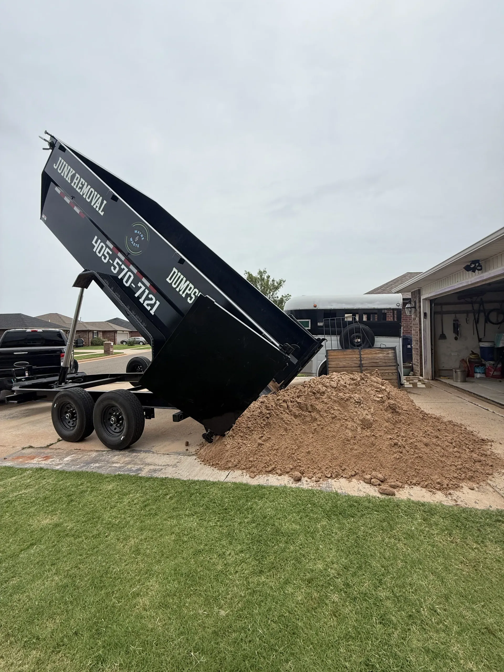 Black dumpster tipped on its side, dumping mulch beside a garage and grassy lawn.