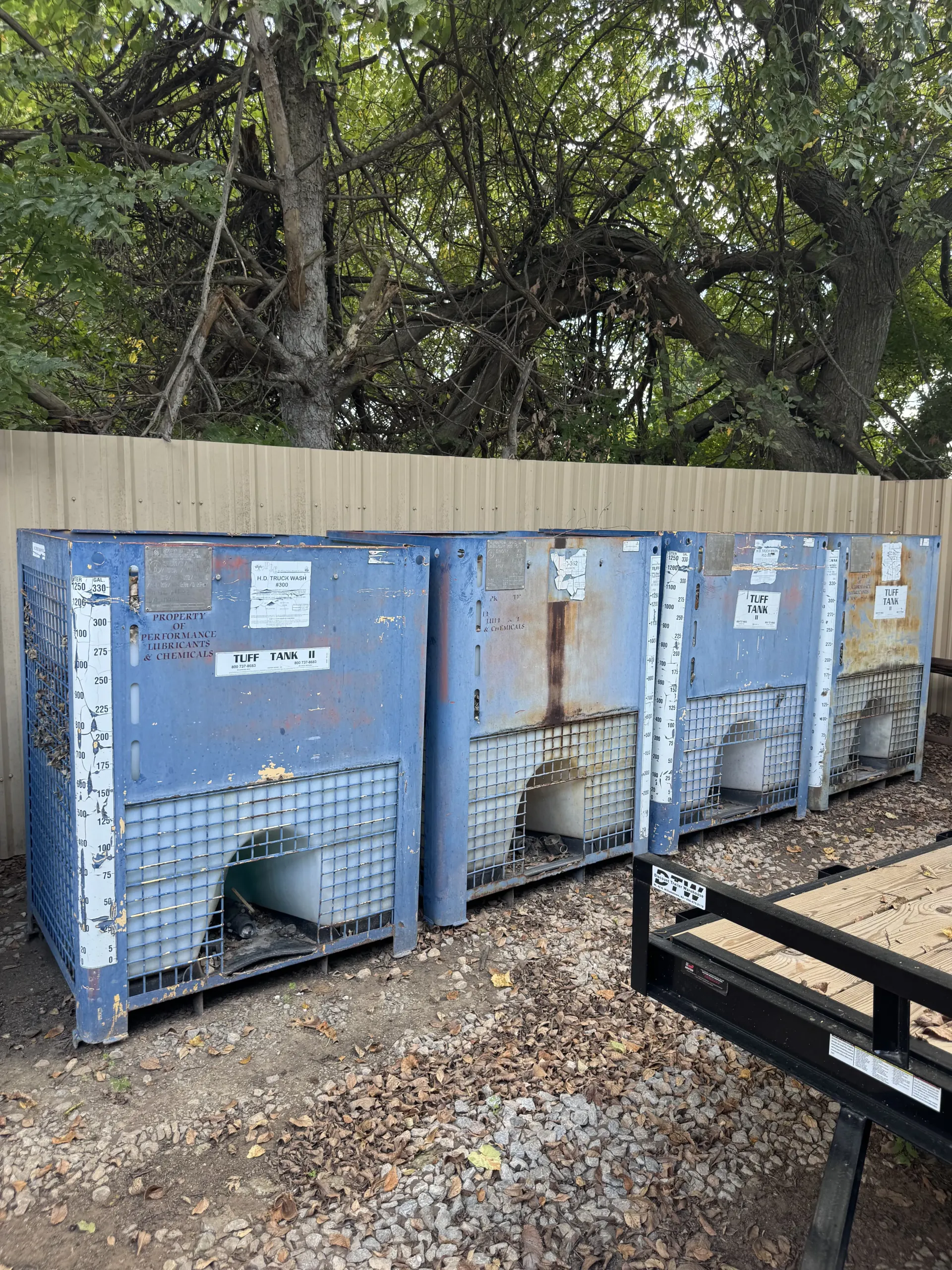 Blue industrial dumpsters against a fence, with trees behind and gravel ground in front.