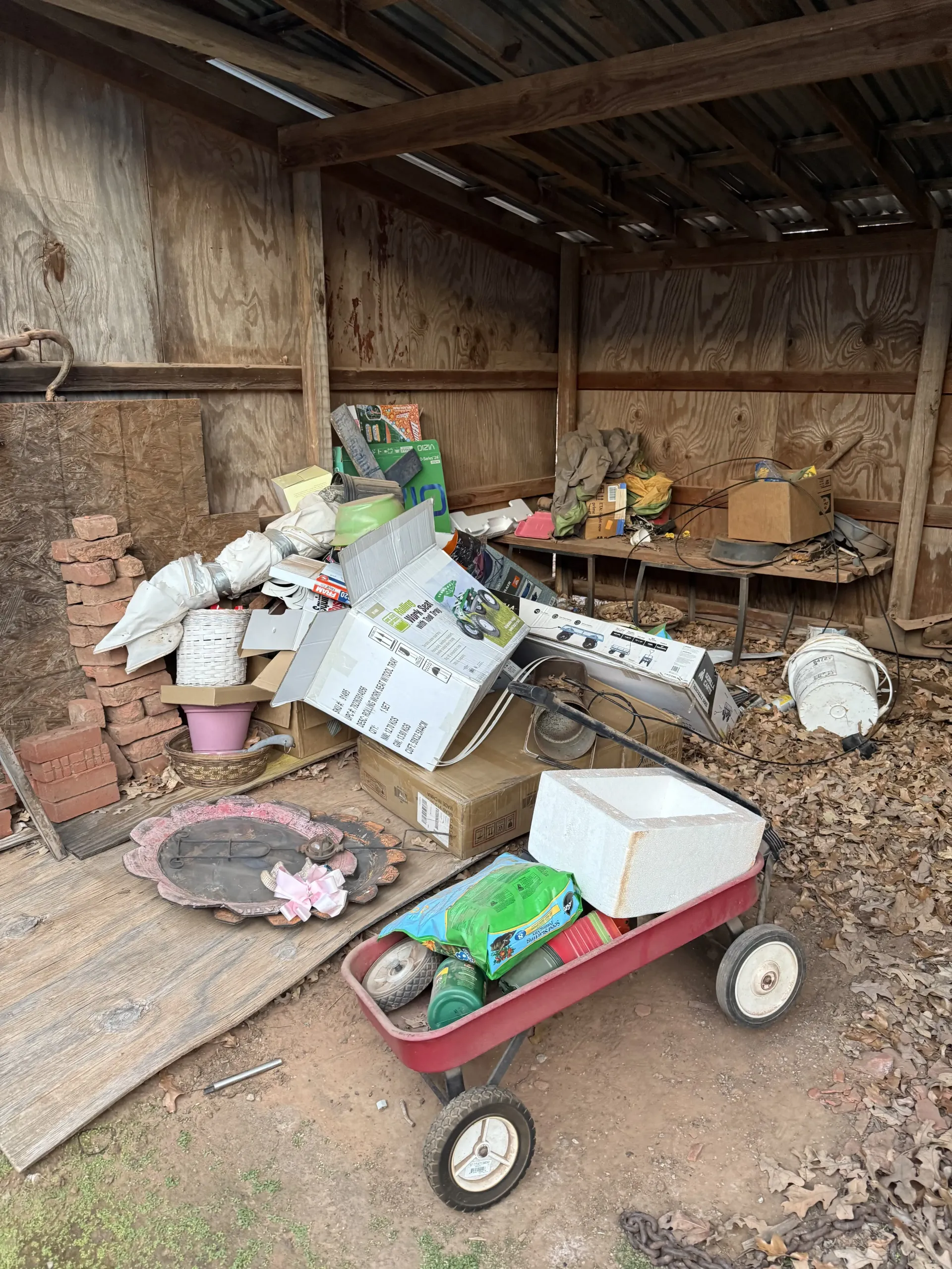 Cluttered barn with scattered tools, leaves, and a red wagon filled with gardening supplies.