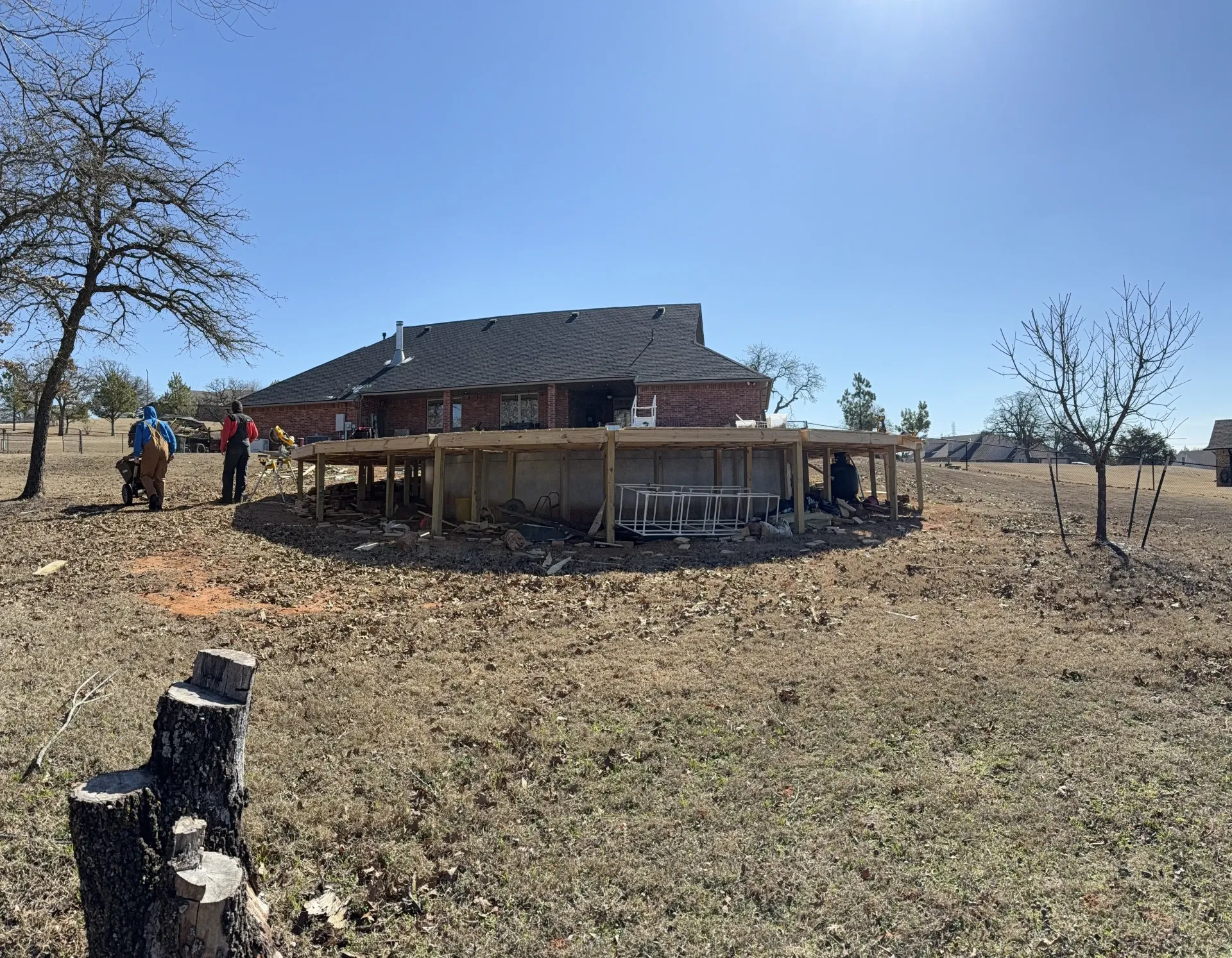 Outdoor scene of a low wooden structure under construction on a dry hillside, with a striped roof and blue sky.
