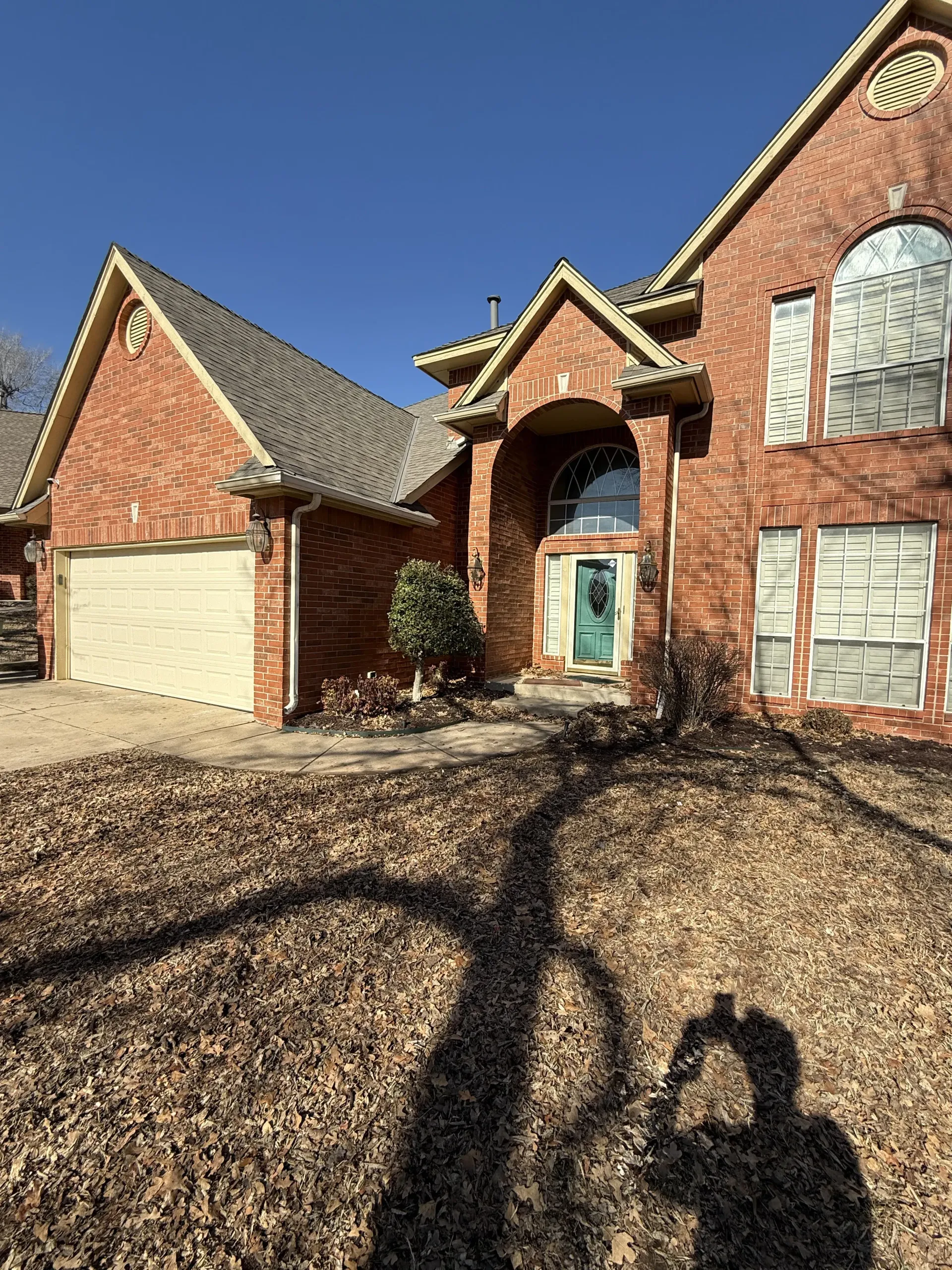 Brick two-story house with arched entry, teal front door, and garage, seen from a leaf-covered yard.