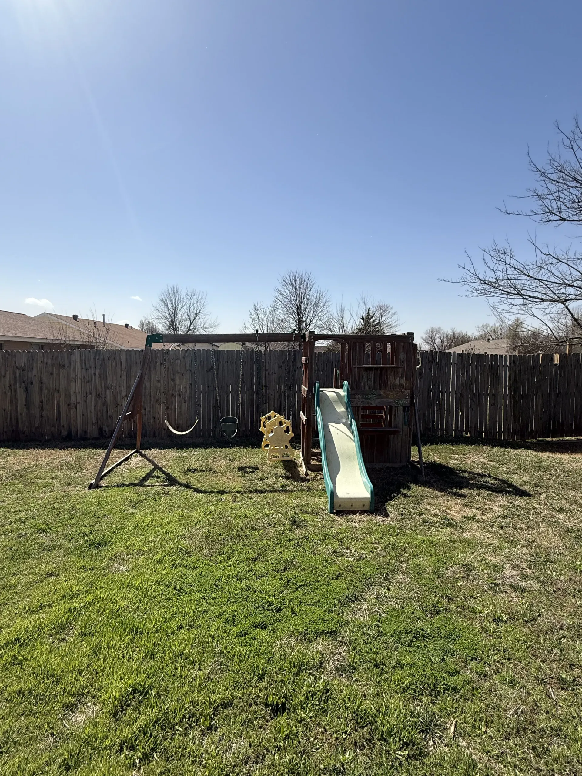 Backyard with a wooden fence and a small green-and-yellow slide under a blue sky.