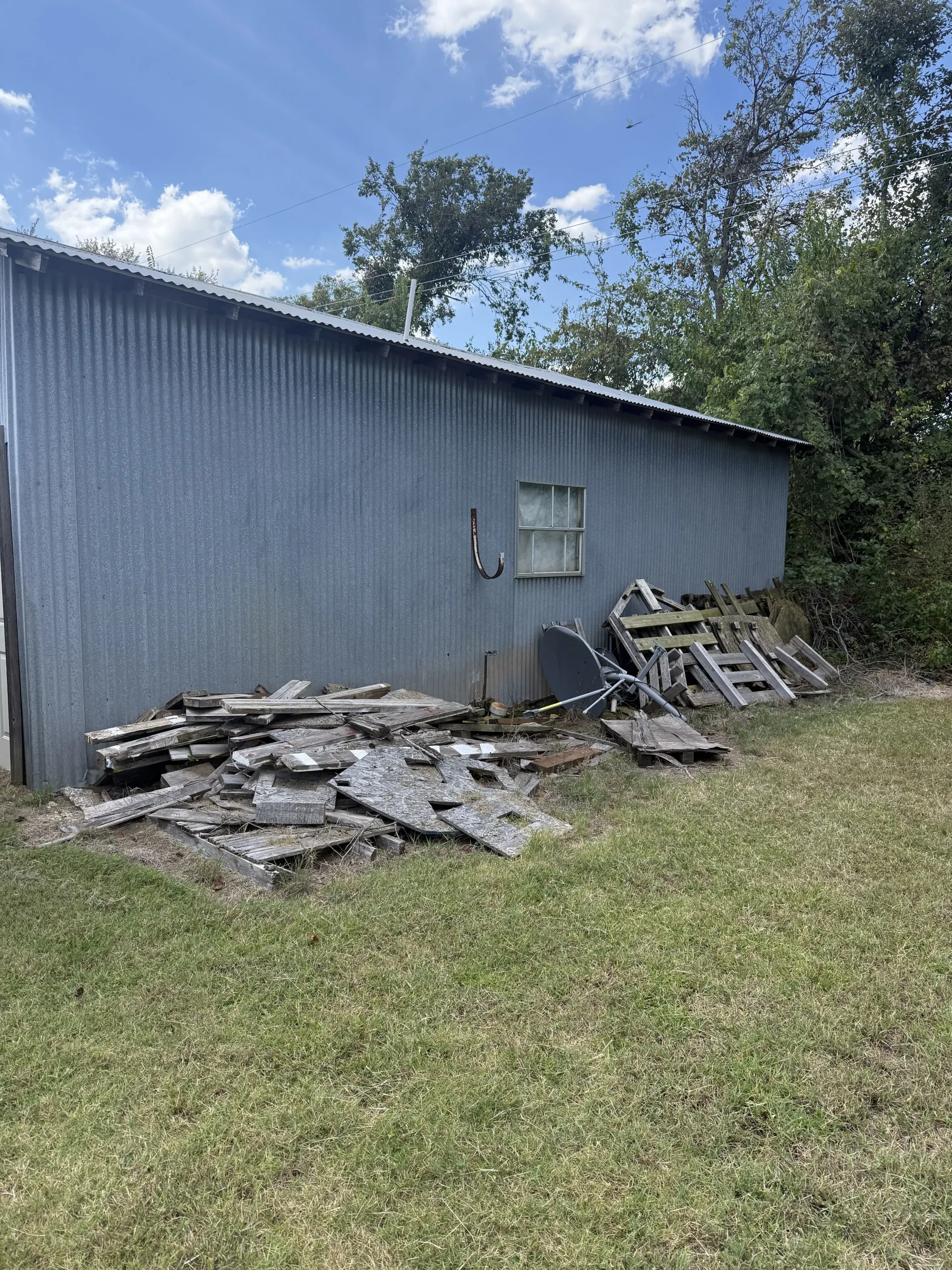 Gray corrugated metal shed with debris piles and stacked materials beside it on a grassy lot under a blue sky