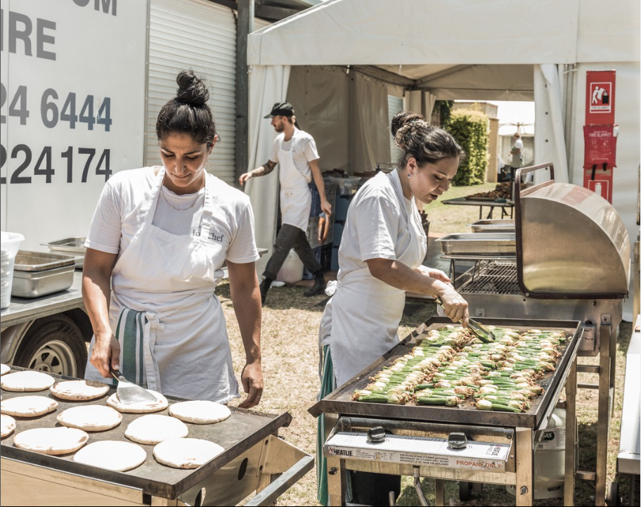 Two women in white aprons are cooking food on a grill.