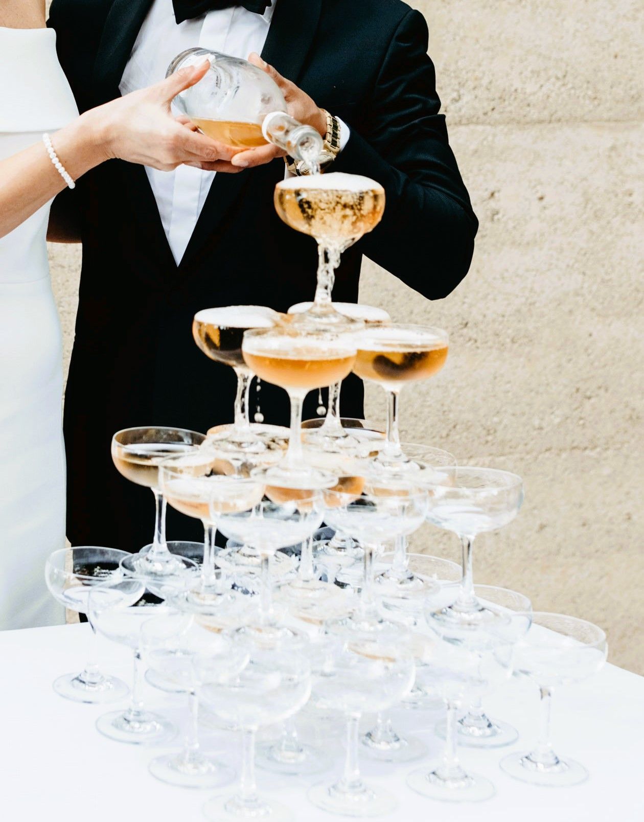 A bride and groom are pouring champagne into a pyramid of champagne glasses.