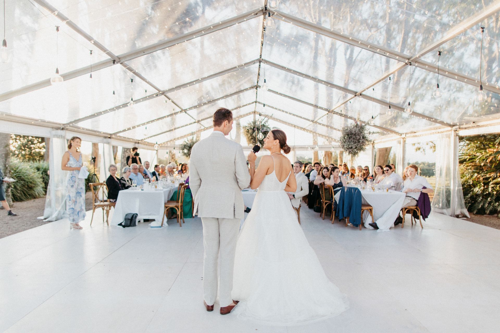 A bride and groom are dancing under a clear tent at their wedding reception.