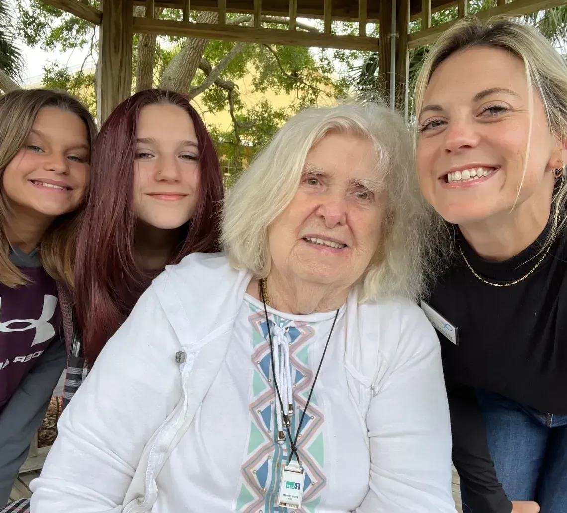 Four smiling people pose together under a gazebo, with an older woman in front and trees behind them.