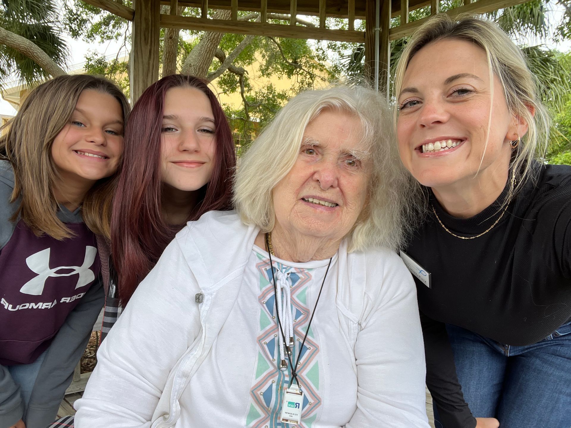 Four smiling people pose together under a gazebo, with an older woman in front and trees behind them.