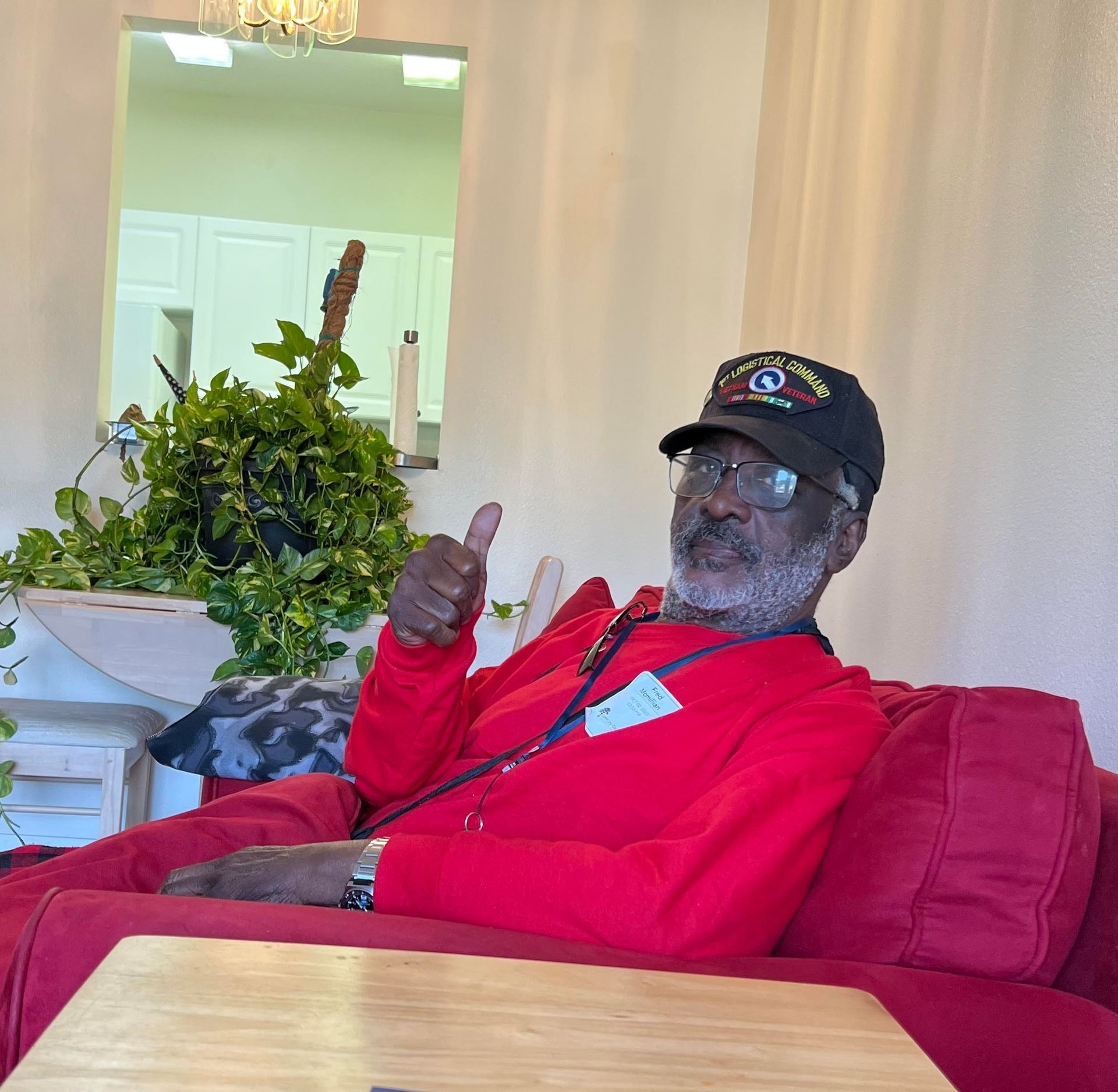 Man in red jacket and cap sitting in a chair, smiling and giving a thumbs-up beside a leafy plant.