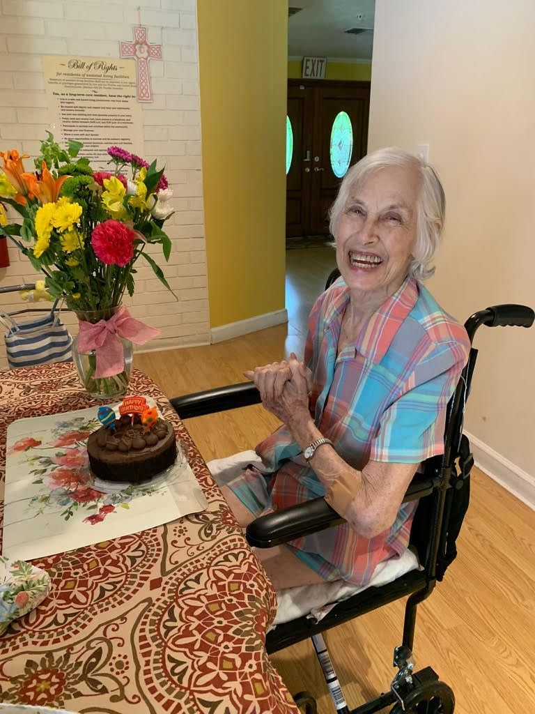 Smiling woman in wheelchair beside birthday cake and flowers at a decorated table, hands clasped in celebration