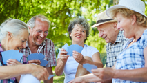 Five seniors smiling and playing cards outdoors under trees on a sunny day