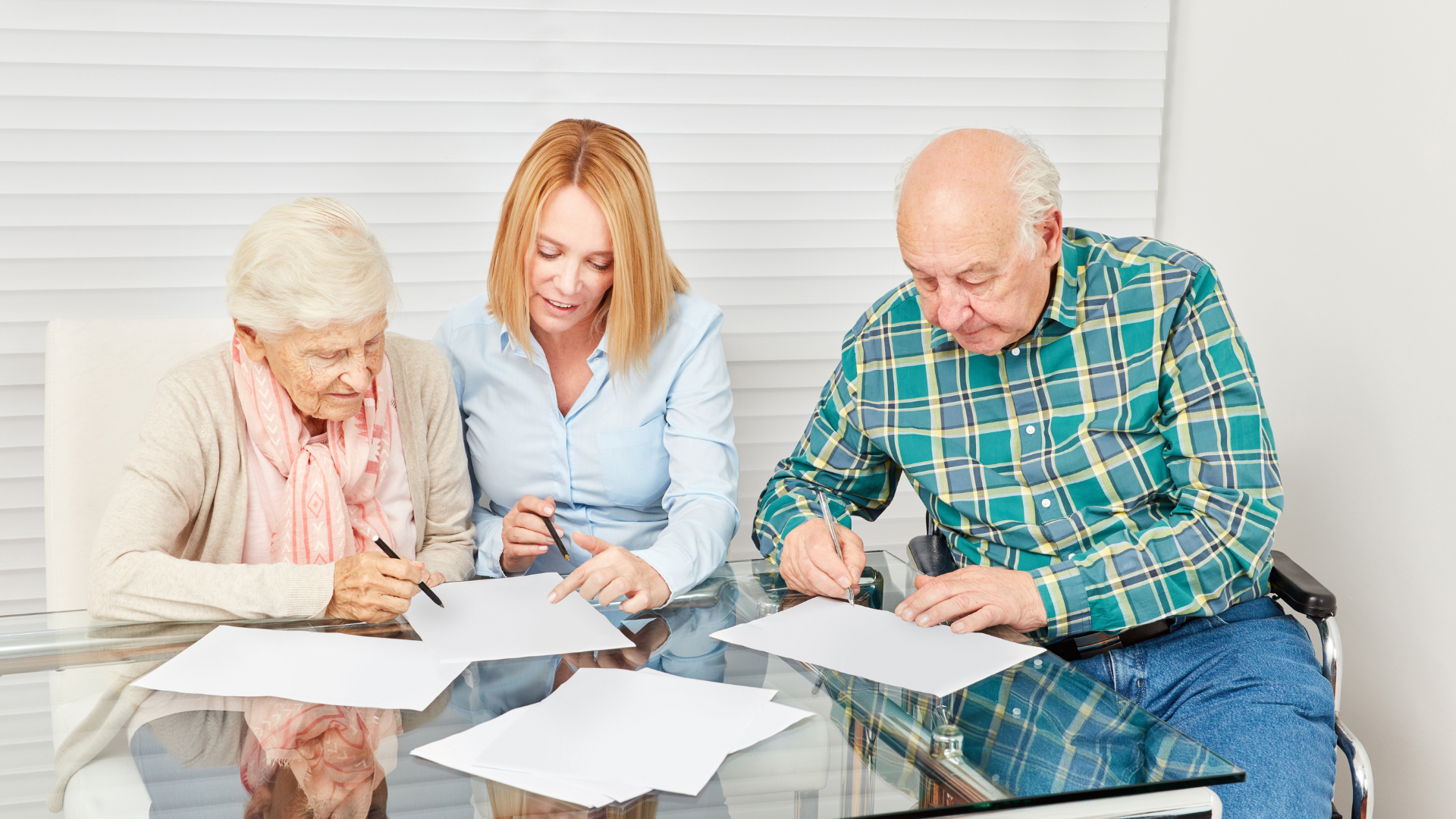 Three people sitting at a glass table reviewing papers and writing notes in a bright room