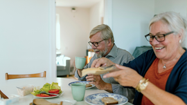 Two people smiling at a dining table with mugs, plates, and a salad in a bright kitchen