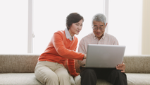 Two people sitting on a couch, smiling and looking at a laptop together.