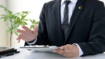 Businessperson in a dark suit gesturing at a desk with a clipboard and plant nearby