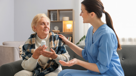Nurse in blue scrubs comforting an elderly patient on a sofa in a living room