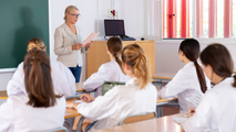 Teacher presenting to seated students in a bright classroom with a laptop and notes.