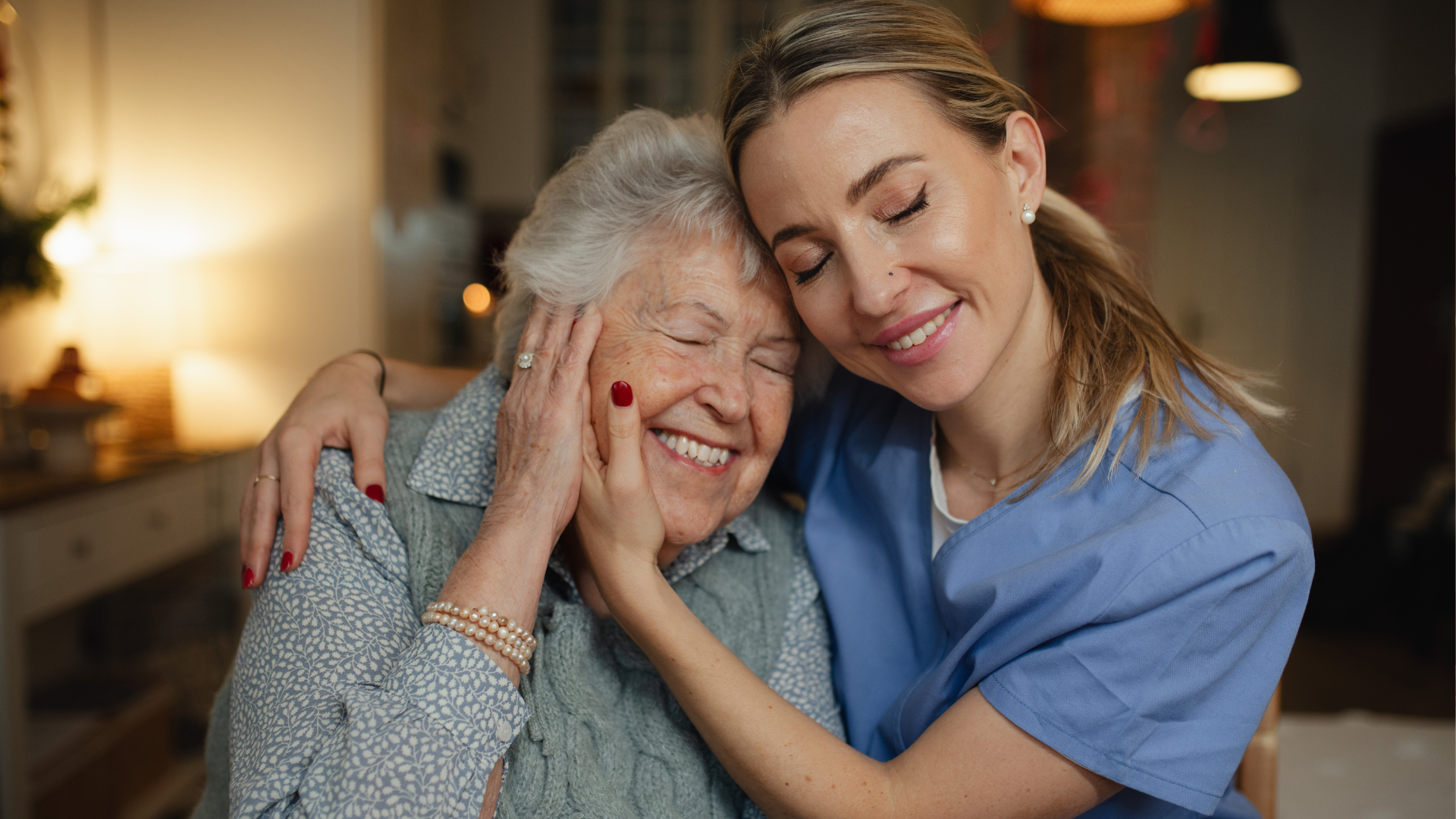 Smiling woman in blue embraces an elderly woman in a cozy living room