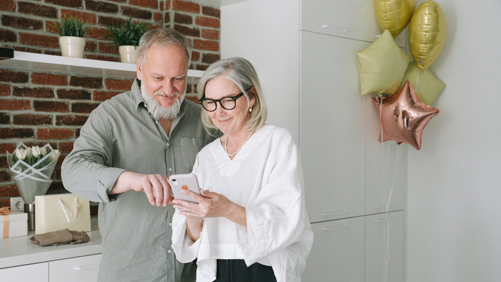 Smiling couple looking at a phone in a kitchen, with balloons and a gift box nearby.