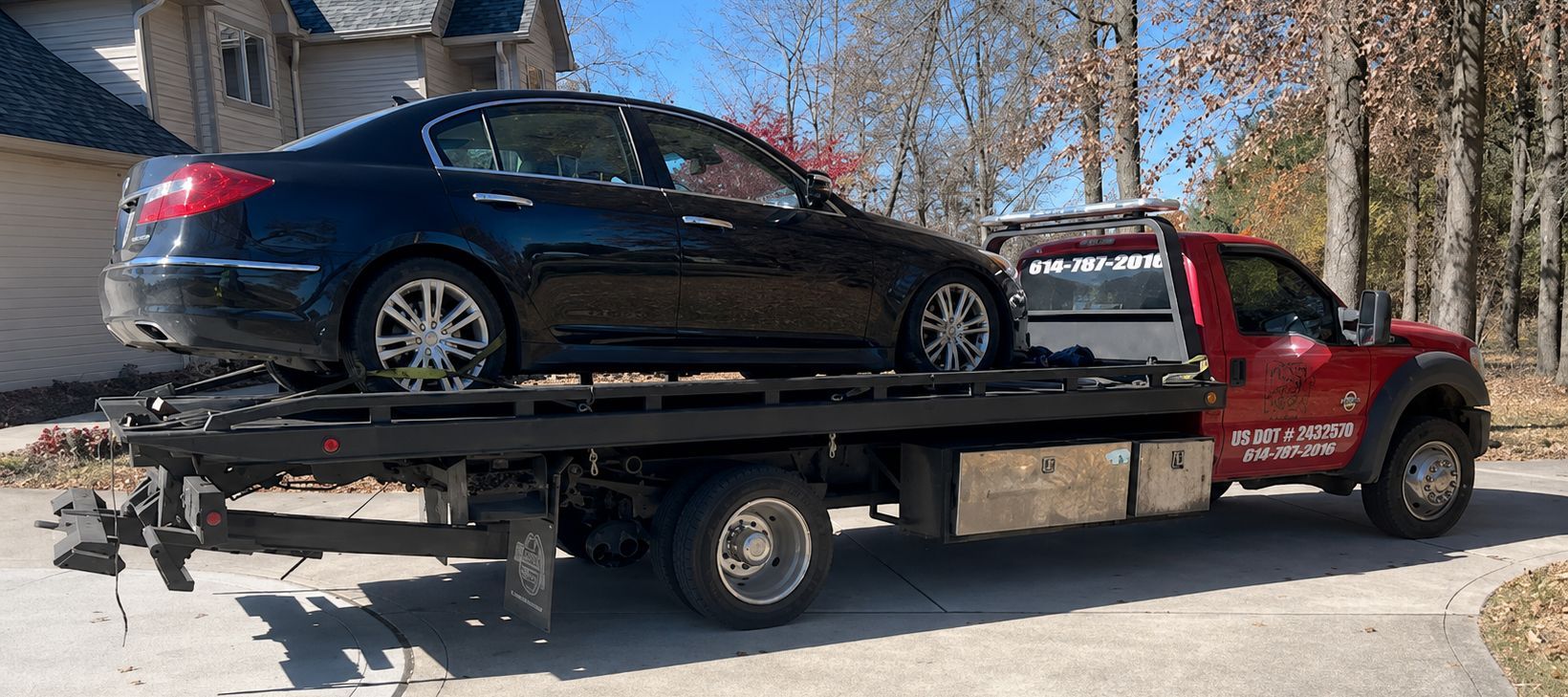 Red tow truck carrying a black sedan on a sunny street with trees in the background