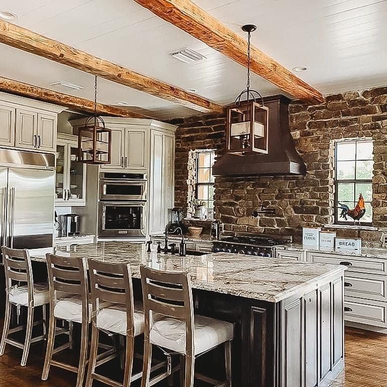 Rustic kitchen with exposed brick wall, granite island, white cabinets, and wooden beams.