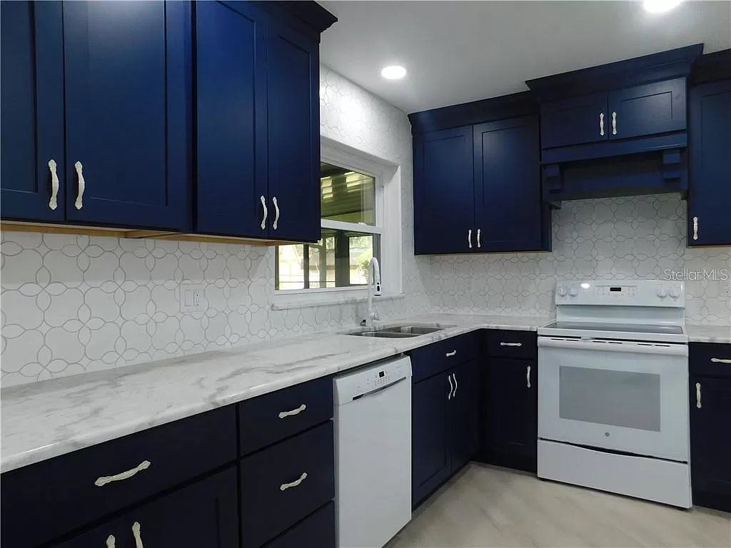 Blue kitchen cabinets, white countertop, white backsplash, white appliances, and a window.