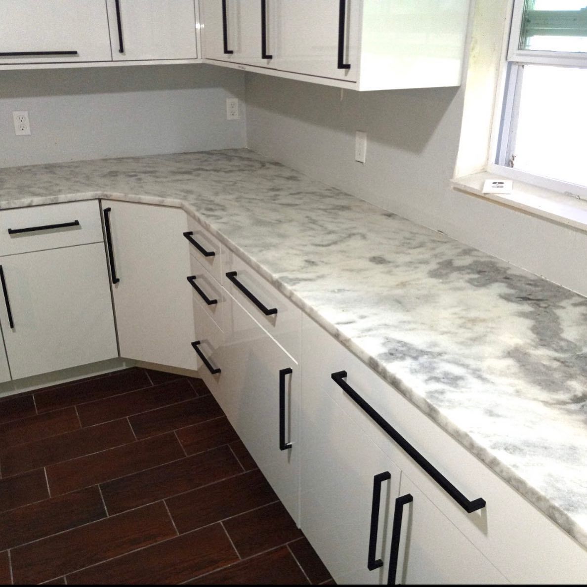 White kitchen cabinets with black handles, marble countertops, and dark wood floors.