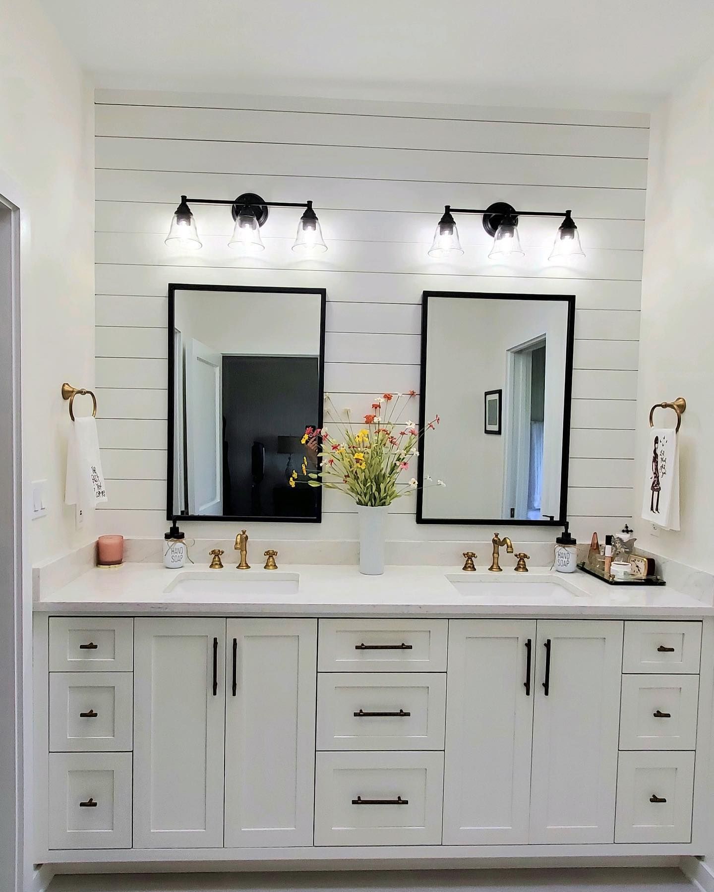 White bathroom with double vanity, two black-framed mirrors, and shiplap wall. Black light fixtures and gold accents.
