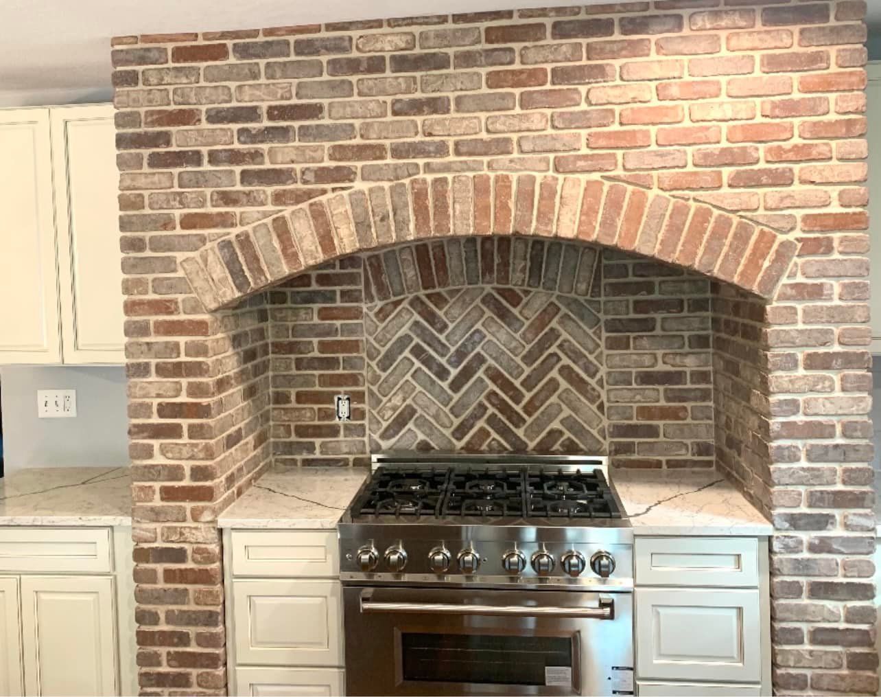 Brick-walled kitchen with stove. Herringbone tile backsplash. Cream cabinets and stainless steel appliances.