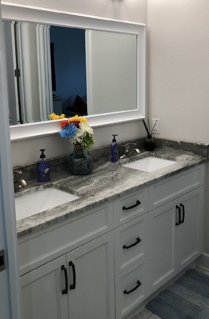 White bathroom vanity with dual sinks, gray countertop, large mirror, and black hardware.