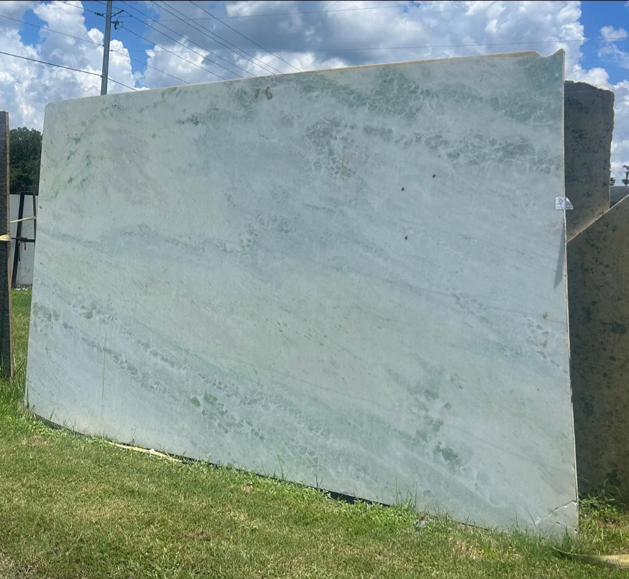Large slab of light blue-green marble, leaning on grass, under a bright, cloudy sky.