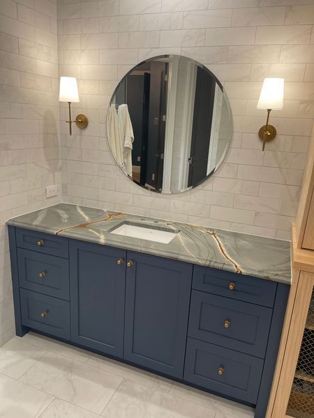 Bathroom vanity with blue cabinets, gray countertop, round mirror, and wall sconces.