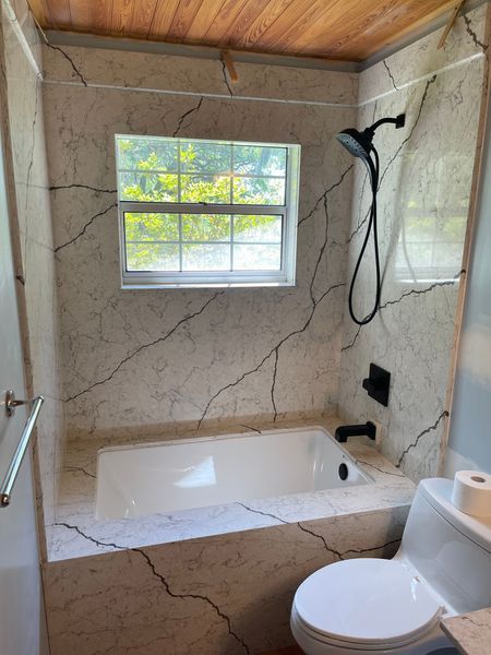 Bathroom with a tub and shower, featuring marble-like walls and a window overlooking greenery. Black fixtures.
