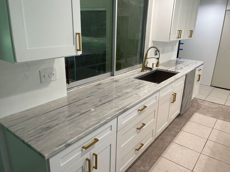 White kitchen cabinets with gray countertops, gold hardware, and a dark sink.